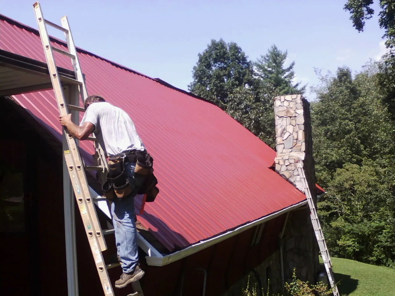 Roofer on a ladder beside a red metal roof and stone chimney — Soffit & Fascia Repair by Roofing Brothers in Warner Springs